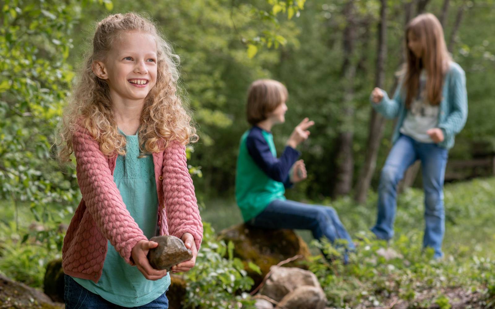 A smiling girl in nature holding a rock and two children playing in the background