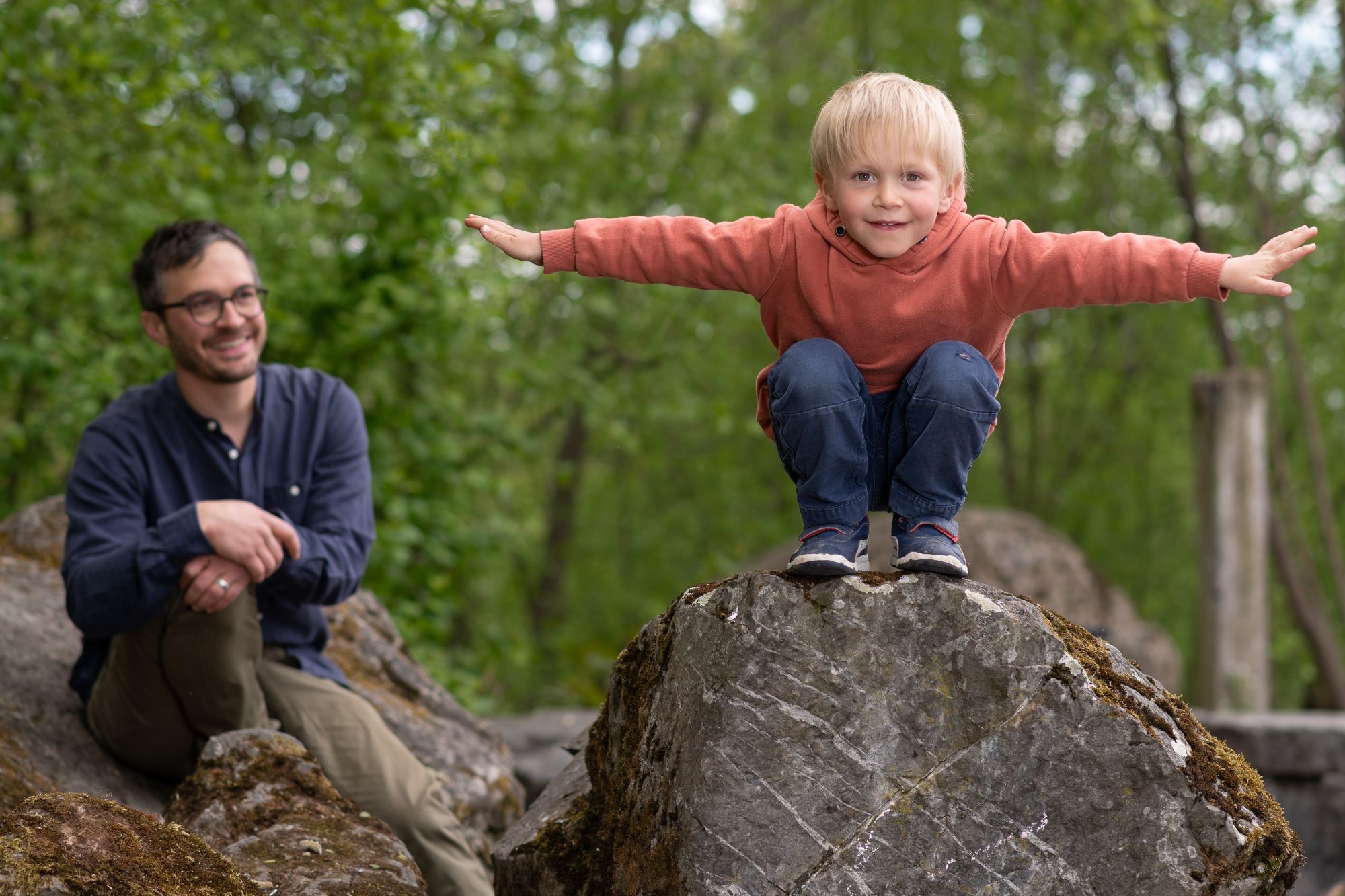 A toddler balancing on a tree trunk and his father smiling in the background