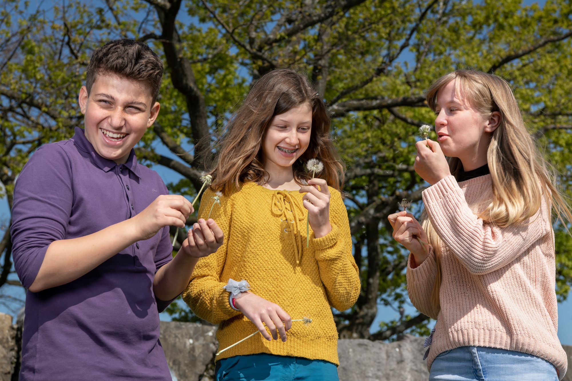 Three teenagers in nature laughing and blowing dandelions