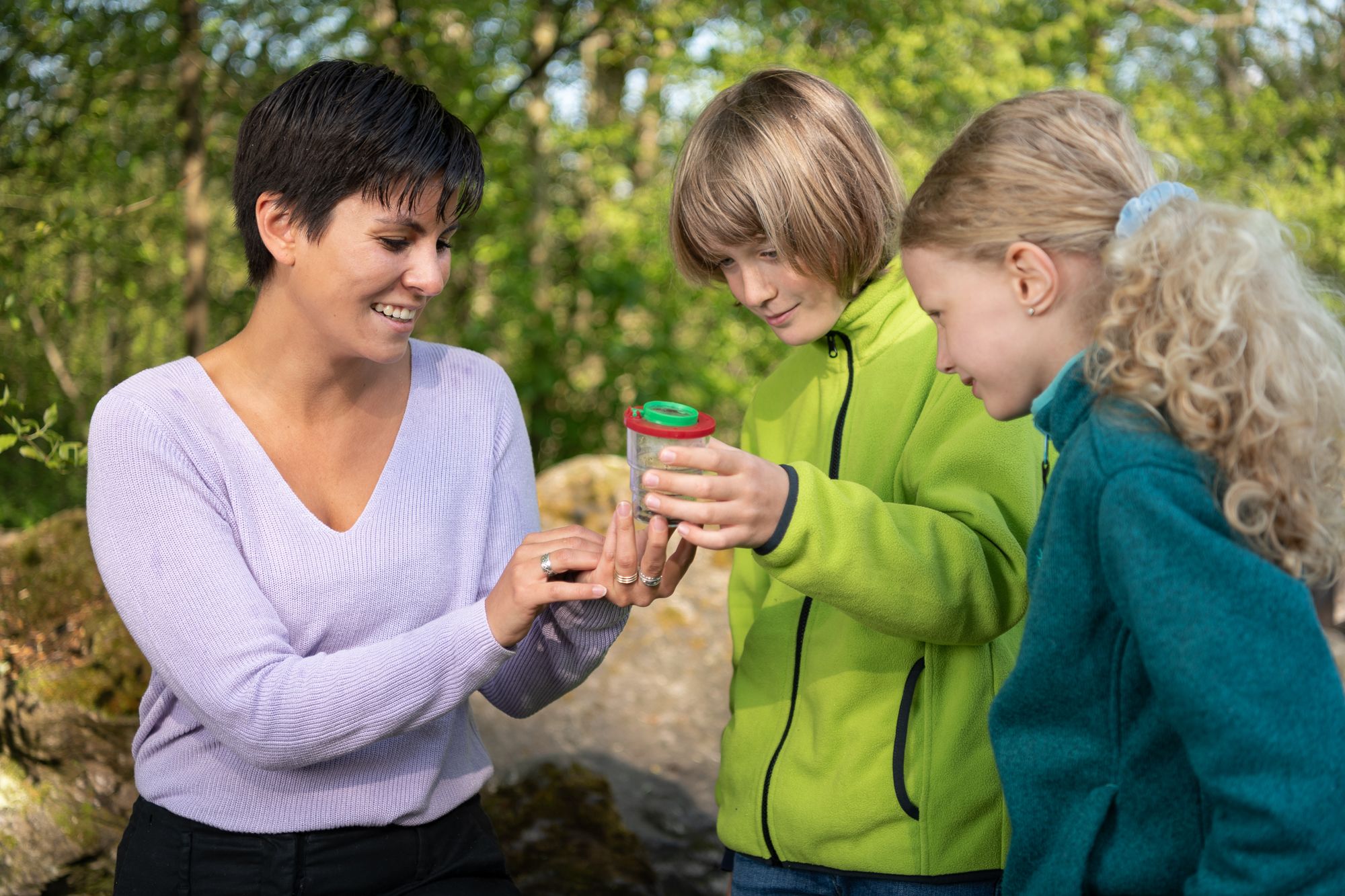 A teacher with two students in nature looking at a newt with a cup magnifier