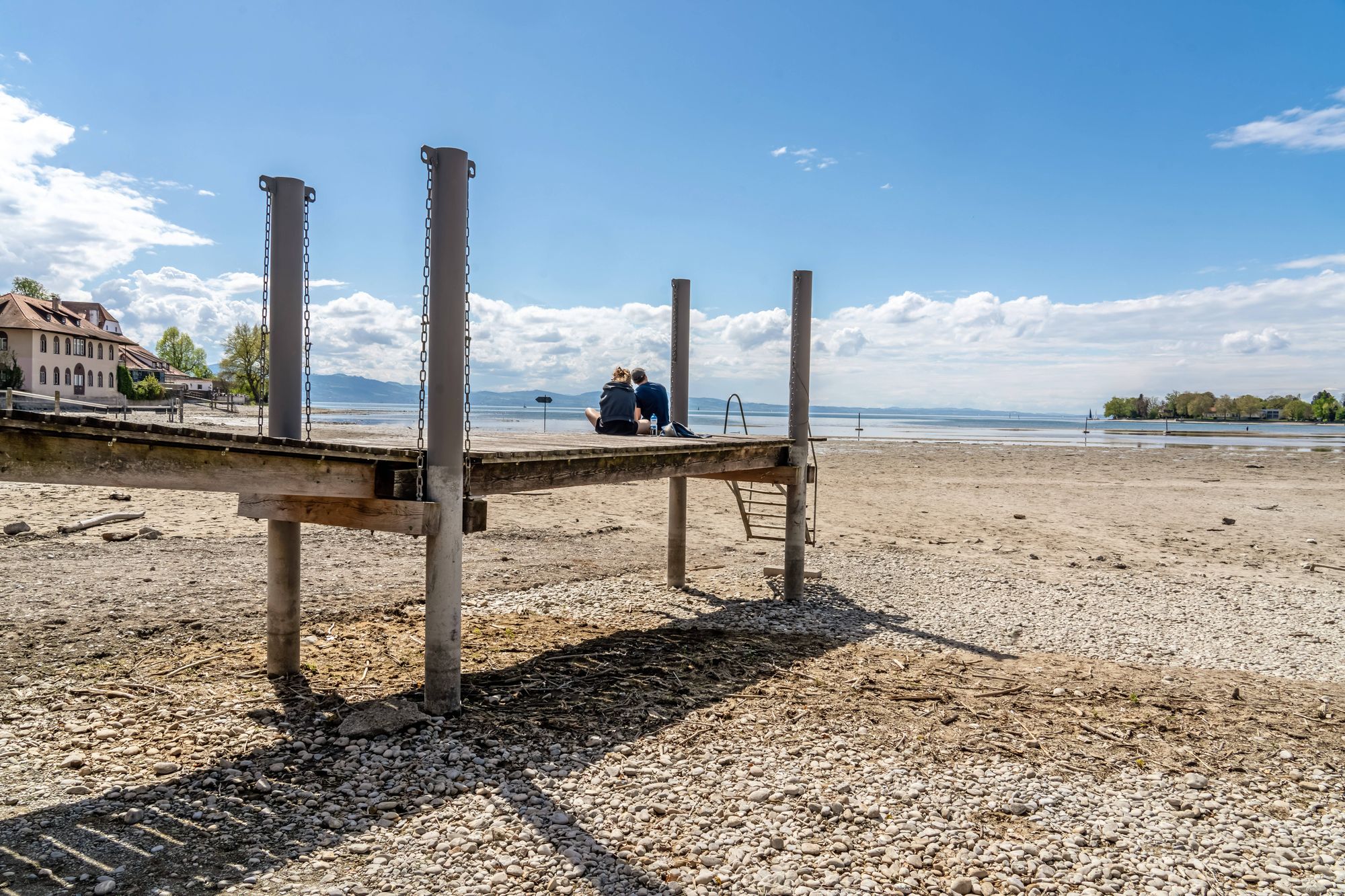 two people sitting on a jetty at lake constance with no water at the shores