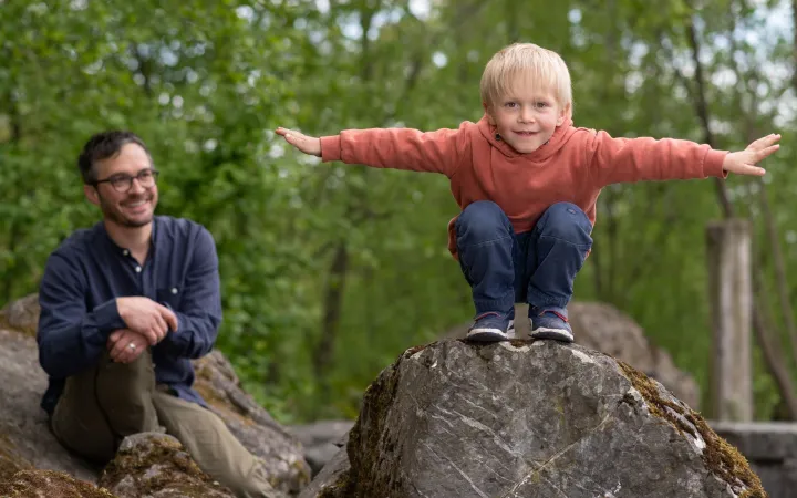 A toddler balancing on a tree trunk and his father smiling in the background