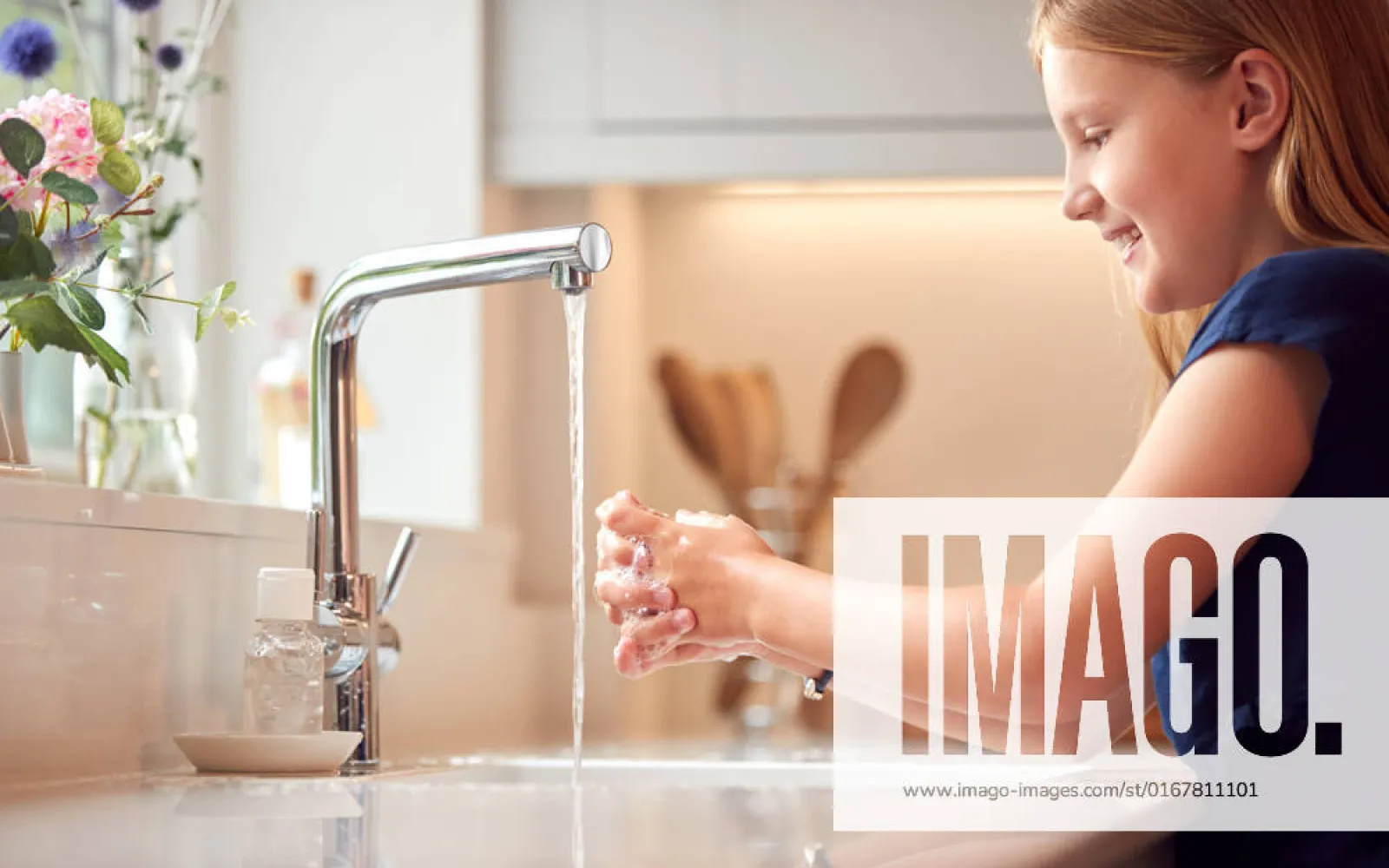 Ein Mädchen wäscht sich in einer modernen Küche die Hände unter einem laufenden Wasserhahn. Im Vordergrund stehen eine Seifenschale und ein Seifenspender, im Hintergrund sind Blumen und Küchenutensilien zu sehen.