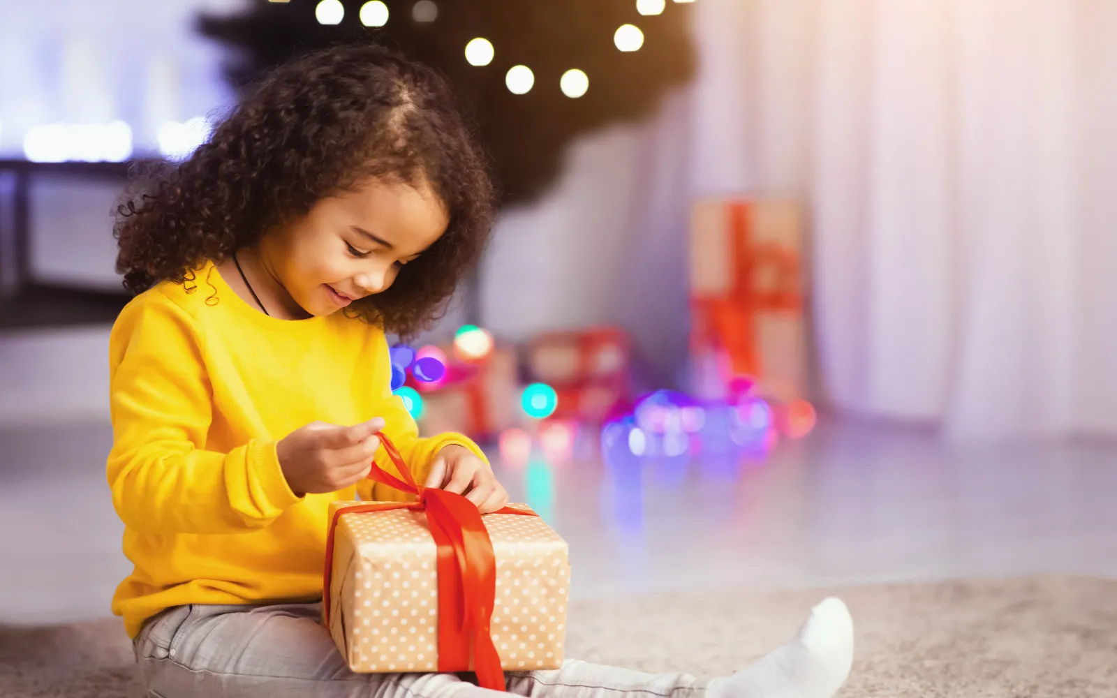 Child sitting in front of a decorated Christmas tree, opening a present with a red ribbon.