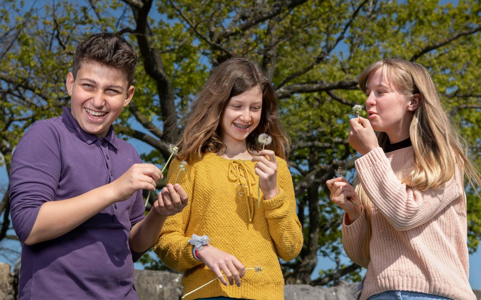 Three teenagers in nature laughing and blowing dandelions