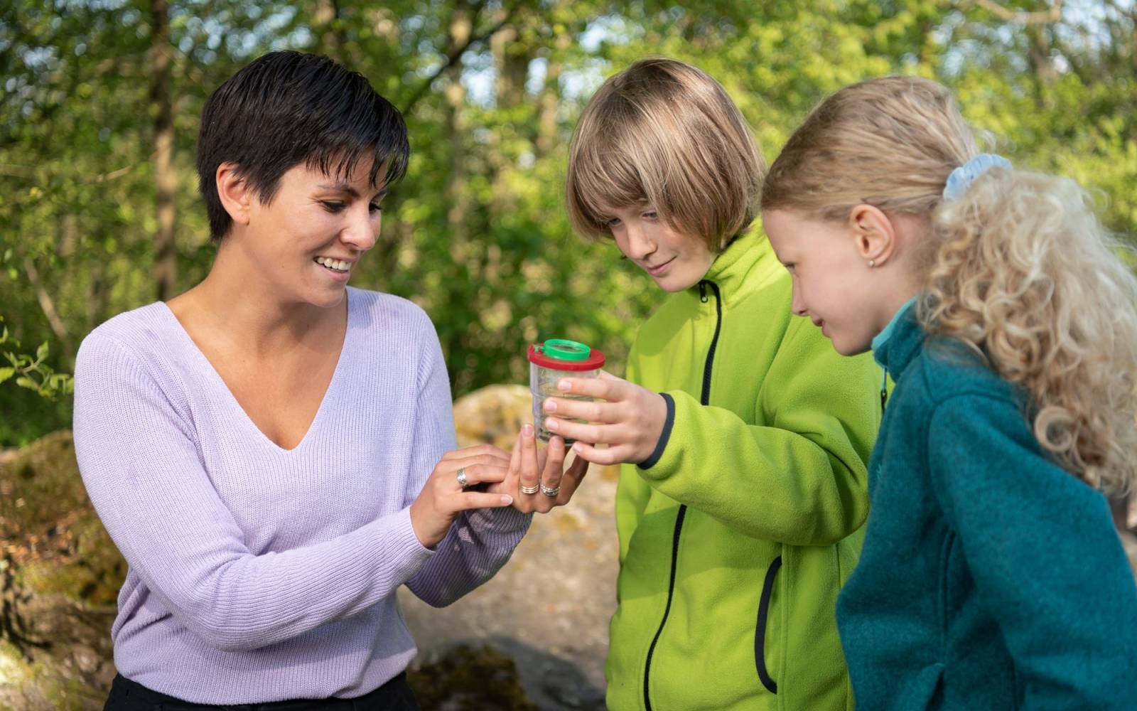 A teacher with two students in nature looking at a newt with a cup magnifier