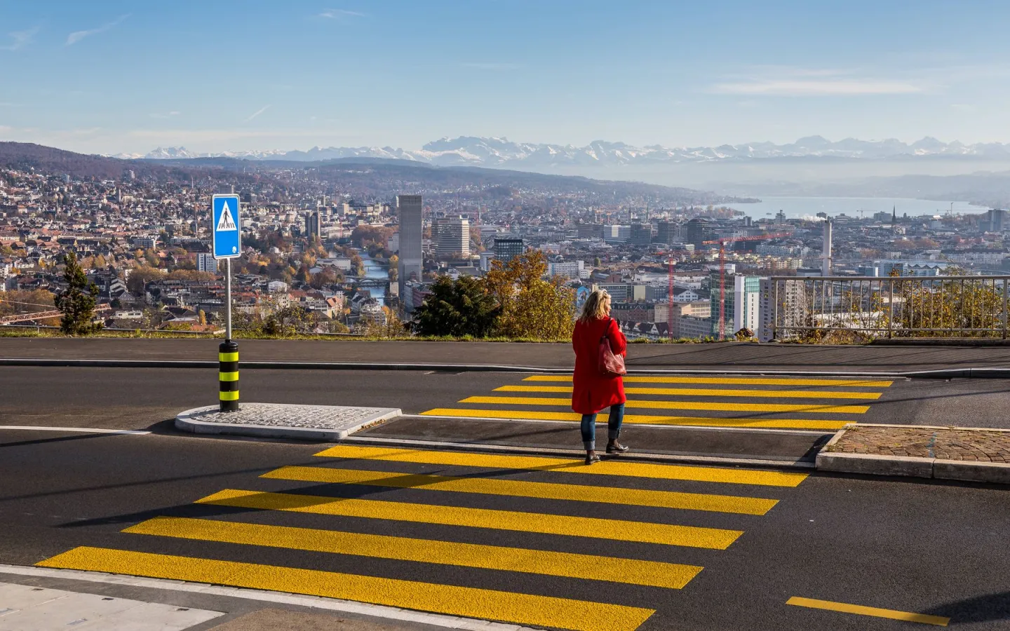 Frau mit rotem Mantel auf Zebrastreifen, im Hintergrund die Stadt Zürich