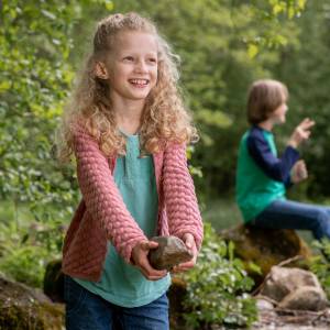 A smiling girl in nature holding a rock and two children playing in the background